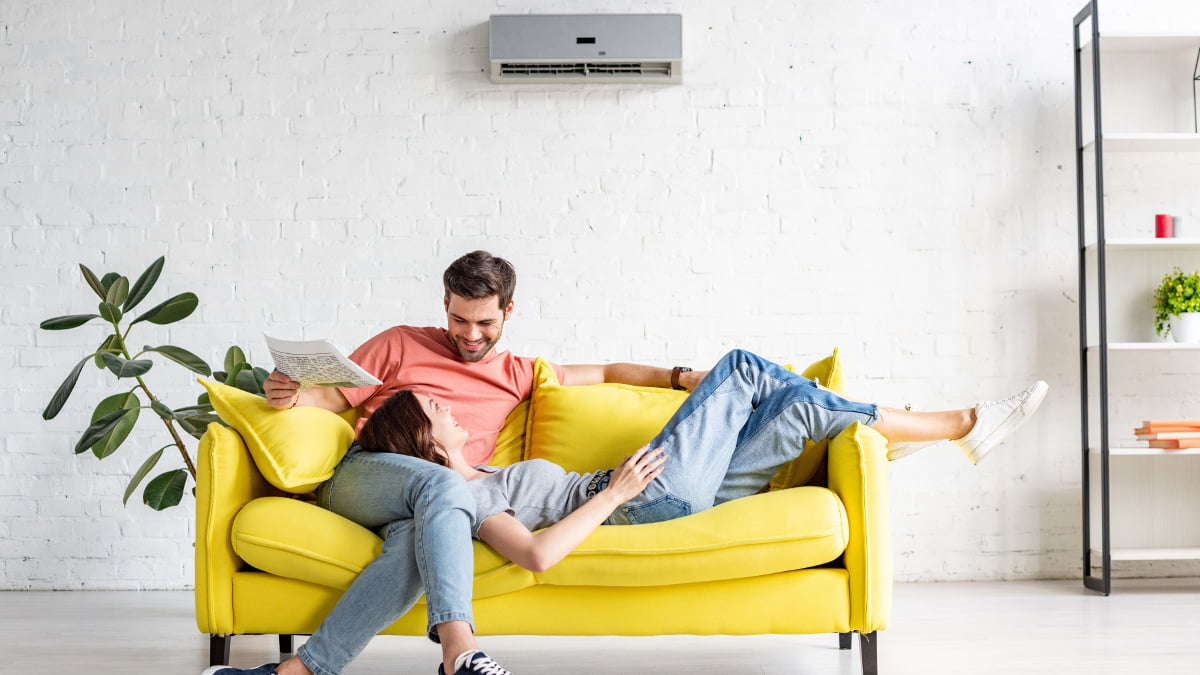 Smiling couple sit on a sofa underneath an air conditioning unit