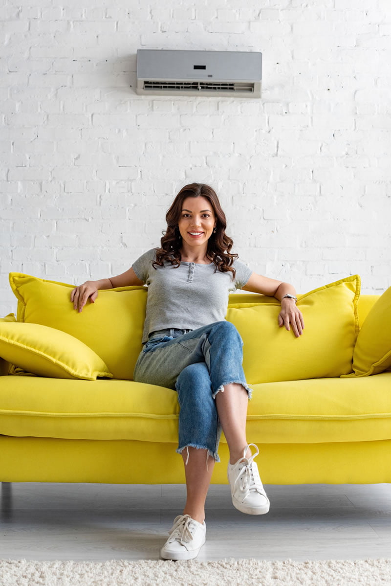 800-air-con Happy woman sat on yellow sofa underneath wall mounted air con unit
