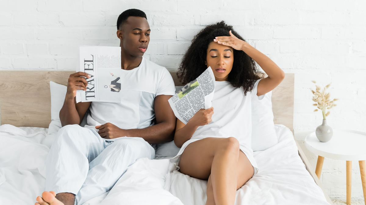 Couple sat on bed fanning themselves with paper to keep cool